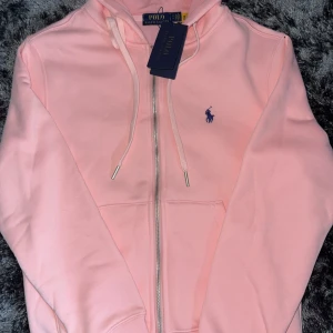 Rosa zip hoodie från Polo Ralph Lauren - Snygg ljusrosa hoodie från Polo Ralph Lauren med blå broderad logga på bröstet. Tröjan har huva med snörning, dragkedja framtill och två fickor. Mjuk bomullskvalitet som känns skön mot huden. Perfekt för chill dagar och streetstyle.