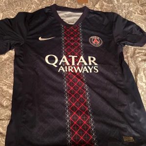PSG matchtröja Nike Qatar Airways - Paris Saint-Germain fotbollströja från Nike med Qatar Airways-tryck. Mörkblå bas med röd och vit geometrisk rand mitt på bröstet, PSG-emblem och Nike-logga. Tillverkad i lätt och ventilerande material, perfekt för match eller träning.