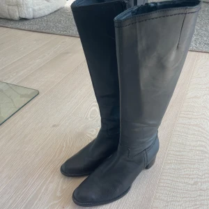 Svarta höga stövlar i skinn - Säljer ett par svarta, klassiska höga stövlar från Billi Bi med rund tå och låg klack. De är tillverkade i mjukt skinn och har dragkedja på insidan för enkel på- och avtagning. Perfekt till jeans eller kjol för en stilren look.