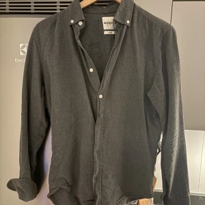 Mörkgrå skjorta från Dobber - Snygg mörkgrå skjorta från Dobber i storlek small. Skjortan har klassisk krage, vita knappar och långa ärmar. Materialet känns mjukt och är troligtvis bomull, perfekt för en chill och stilren look. Passar dig som gillar enkel och clean stil.