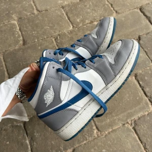 Nike Air Jordan 1 Mid grå/blå - Säljer ett par Nike Air Jordan 1 Mid i storlek 38. Skorna har en clean färgkombo med grått och vitt skinn samt blå detaljer och snörning. Klassisk Jordan-logga på sidan och blå swoosh. Perfekta för dig som vill ha en ikonisk sneaker med snygg streetstyle-vibe. Nypris 1800kr                                                   Super nice Jordan Mids in the color True Blue. Never worn, bought in London at JD Sports. Size 38 (UK 5). Unisex – suitable for both girls and boys.