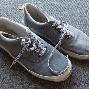 Grå sneakers med textade snören - Grå sneakers i canvas med vita sulor och snörning. Skosnörena är vita med svart text 'SLEEP PLAY REPEAT' tryckt längs hela snöret. Skorna har rund tå och en klassisk, enkel design som passar till det mesta.