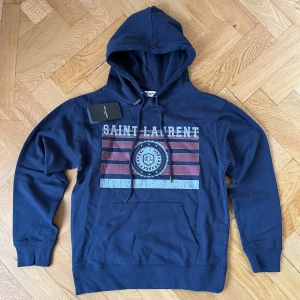 Marinblå Saint Laurent hoodie - Marinblå hoodie från Saint Laurent med tryckt logga och grafiskt mönster i vitt och rött framtill. Klassisk känguruficka, snörning i huvan och ribbade muddar. Perfekt för dig som vill ha en snygg streetstyle-look.