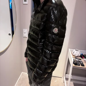 Svart pufferjacka från Moncler - Säljer en svart, glansig pufferjacka från Moncler med klassisk logga på ärmen. Jackan har dragkedja framtill, ståkrage och quiltad design. Perfekt för dig som vill ha en snygg och varm jacka med streetkänsla.