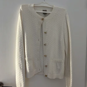 Vit kofta med guldknappar från H&M - Säljer en stilren vit kofta från H&M i storlek S. Koftan är stickad med diskret struktur, har två fickor framtill och snygga guldiga knappar. Perfekt att slänga över en t-shirt eller klänning för en clean look.