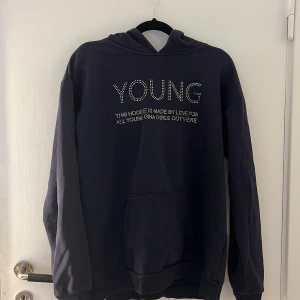 Mörkblå hoodie med strass från Gina Tricot - Snygg mörkblå hoodie från Gina Tricot med texten 'YOUNG' i strass på bröstet. Klassisk känguruficka framtill och huva. Perfekt för dig som gillar en chill och trendig stil.