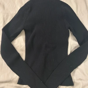 Svart ribbad tröja från Lindex - Svart tröja från Lindex i storlek S. Tröjan är ribbstickad med snygga detaljer längs sidorna och bröstkorgen och har lång ärm. Den är tight i passformen och har en hög krage.