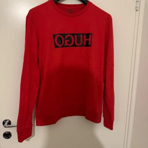 Röd långärmad tröja från Hugo - Snygg röd långärmad tröja från Hugo med svart logotyptryck på bröstet. Tröjan har rund halsringning och är gjord i mjuk bomull som känns skön mot huden. Perfekt för dig som gillar stilrena och trendiga plagg med tydlig branding.