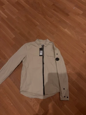 Beige overshirt från C.P. Company - Snygg beige overshirt från C.P. Company med klassisk krage, svart dragkedja och ikonisk linsdetalj på ärmen. Jackan har en bröstficka med knapp och är tillverkad i ett lätt och stilrent material. Perfekt för lager-på-lager och streetwear-vibe.