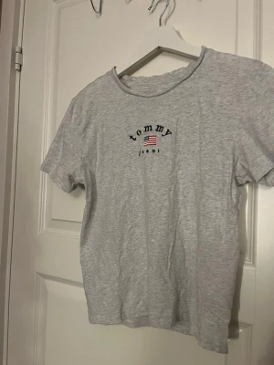 Grå t-shirt Tommy Jeans  - Säljer en grå t-shirt från Tommy Jeans i storlek S. T-shirten har korta ärmar och är gjord i mjuk bomull. På bröstet finns ett broderat Tommy Jeans-tryck med amerikansk flagga och på ena ärmen sitter en liten logotyp. Perfekt för en chill och stilren look.
