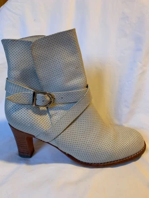 Beige perforerade stövletter med klack - Snygga benvita/ljust beige stövletter med perforerat mönster och rund tå. Skorna har en bekvämt hög klack på 7,5 cm i trälook och ett dekorativt spänne med omlottrem runt ankeln. Tillverkade i skinn. Använda men väl omhändertagna. Smärre defekter och slitage finns men inget som stör, helhetsintrycket är mycket gott. 