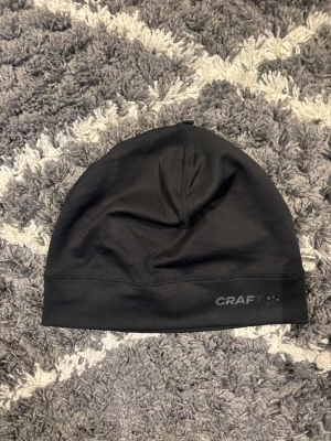 Svart mössa från Craft - Svart mössa från Craft i slät och stretchig polyester. Enkel design med diskret logga framtill. Perfekt för dig som gillar sportig stil och vill ha något som sitter skönt på huvudet under kyliga dagar.