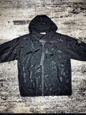 Svart vindjacka från Stone Island - Svart vindjacka från Stone Island med huva och dragkedja framtill. Jackan har två snedställda fickor med dragkedja och den klassiska Stone Island-loggan på ärmen. Materialet är polyester och jackan har en clean, urban look som passar streetwear-stilen.