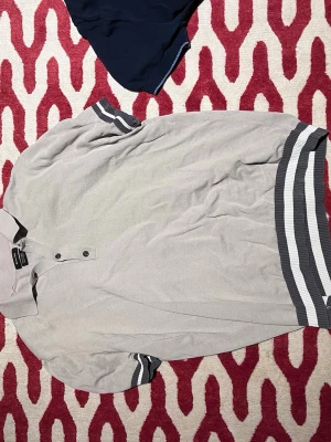 Beige piké med randiga muddar - Säljer en beige piké från Massimo dutti med grå och vita ränder på ärmslut och nederkant. Klassisk krage och två knappar framtill. Perfekt till jeans eller shorts för en clean look.