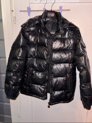 Svart pufferjacka med huva från Cavaliere - Svart pufferjacka  med glansig finish, huva och praktiska fickor med dragkedja och knapp. Jackan har långärmade ärmar och quiltad design som ger en cool och modern look. Perfekt för kalla dagar när du vill vara både varm och stilren.  Köptes till dottern förra vintern så använd endast ett par månader.  Oklart vilket märke det är o det finns inga lappar mer än tvättanvisningar. Köptes på nätet för 899kr. Som ny!  Innerficka, knappar i ärmslut, dragkedja för fickorna på framsidan, luva. Varm jacka