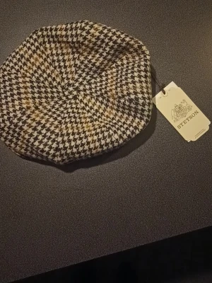 Rutig keps Stetson brun/beige - Säljer en rutig keps från Stetson i storlek 59/L. Västen har ett klassiskt rutmönster i brunt, beige och svart, med en mjuk och lyxig känsla. Materialet är ull och insidan är fodrad med sidenliknande tyg. Perfekt för dig som gillar vintagevibbar och tidlös stil.