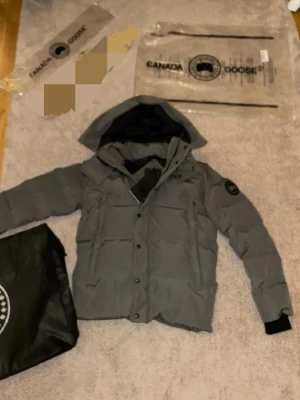 Helt ny oanvänd grå pufferjacka från Canada Goose i storlek och passform L-XL - Säljer en grå pufferjacka från Canada Goose med huva och svarta detaljer. Jackan har knappar och dragkedja framtill, samt ett snyggt märke på ärmen. Perfekt för kalla dagar och har en clean, modern look som funkar till streetwear.