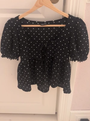 Svart prickig blus från Gina Tricot - Supersöt svart blus med vita prickar från Gina Tricot. Modellen har puffiga korta ärmar, volang nedtill och två knytband framtill. Materialet är lätt och luftigt, perfekt för varmare dagar. Passformen är normal och blusen har en feminin, flowy siluett.
