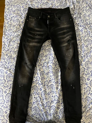 Svarta skinny jeans från Dsquared2 - Säljer ett par svarta skinny jeans från Dsquared2 med slitningar och coola färgstänk på benen. Jeansen har klassisk femficksdesign, knappgylf och en stor vit patch bak i midjan. Materialet är stretchigt jeans och passformen är tight. Använt ett fåtal gånger
