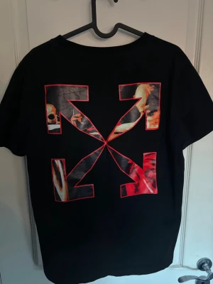 Svart Off-White t-shirt med tryck - Säljer en svart t-shirt från Off-White med stort grafiskt tryck i rött, orange och vitt på ryggen och en konstnärlig print med skelett och händer på bröstet. T-shirten har korta ärmar och är gjord i mjuk bomull. Perfekt för dig som gillar streetwear och unika prints.