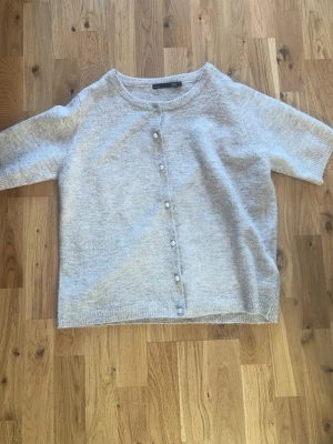 Beige kofta med pärlknappar - Mysig beige  kofta med korta ärmar och pärlknappar framtill. Koftan har en rund halsringning och är stickad i mjukt material som passar perfekt för lager-på-lager. Enkel och stilren design som funkar till många olika looks.