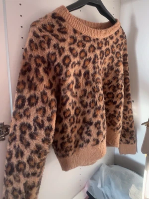 Leopardmönstrad stickad tröja - Säljer en fluffig stickad tröja med leopardmönster i bruna och beige toner. Tröjan har rund hals och långa ärmar med ribbade muddar. Perfekt för dig som vill ha en cool och mysig look under hösten och vintern.