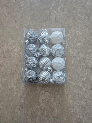 Julkulor - Säljer ett 12-pack julkulor i silver och vitt från Rusta. Kulorna är ca 3,5 cm i diameter och har olika mönster, bland annat glittriga och geometriska former. Perfekt för att ge granen en snygg och modern touch. Materialet är plast.