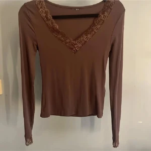 Brun långärmad topp med spets - Säljer en brun långärmad topp med v-ringning och dekorativ spets runt halsringningen och vid ärmsluten. Toppen har en slim passform och är tillverkad i ett mjukt material som känns skönt mot huden. Perfekt att styla med jeans eller kjol.