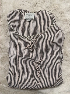 Randig klänning från Masai XXL - Säljer en snygg ärmlös klänning från Masai i storlek XXL. Klänningen har svartvita vertikala ränder och knytning framtill vid halsen. Materialet känns mjukt och luftigt, perfekt för varmare dagar. .