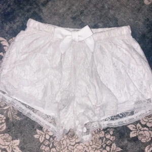 Vita spets-pyjamas shorts med rosett XS - Supersöta vita shorts i spets från C, storlek XS. De har en bred resår i midjan och en stor rosett framtill. Spetsen ger en romantisk vibe och kanten är dekorerad med fin spetsdetalj. Perfekta för varma dagar eller när du vill känna dig extra gullig.