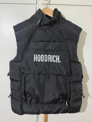 Svart dunväst från Hoodrich - Svart dunväst från Hoodrich med stor vit logga på bröstet. Västen har hög krage med mjukt fleecefoder, justerbara spännen på sidorna och en quiltad design. Perfekt för lager-på-lager och streetwear-stil.