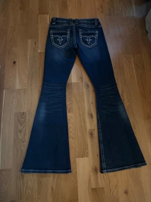 Blå bootcut jeans med slitningar - Säljer ett par mörkblå bootcut jeans med slitningar och snygga detaljer på bakfickorna. Jeansen har kontrastsömmar och en klassisk femficksmodell. Materialet är jeans med en skön stretch och passformen är loose med riktigt utsvängda ben.
