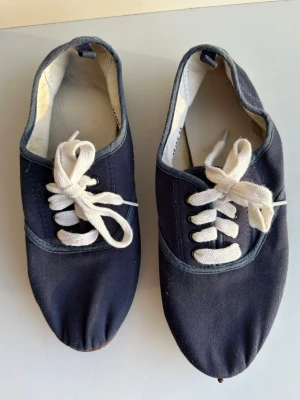 Mörkblå sneakers med vit snörning - Säljer ett par mörkblå sneakers med vit snörning och rund tå. Skorna har en platt sula och är gjorda i bomullstyg med grå kantdetaljer. Perfekta för dig som gillar enkel och klassisk stil. Snygga att matcha med jeans eller shorts.