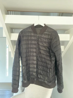 Svart quiltad bomberjacka McKinley - Quiltad svart bomberjacka från McKinley med ribbade muddar och krage. Jackan har dragkedja framtill och två sidofickor med dragkedja. Perfekt för dig som gillar en clean och sportig look. Materialet är syntet och jackan har ett diskret märke på ärmen.