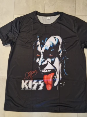 Svart KISS t-shirt med tryck - Cool svart t-shirt med stort tryck av Gene Simmons från KISS på både fram- och baksidan. T-shirten har korta ärmar och är gjord i mjuk polyester som känns skön mot huden. Perfekt för dig som vill sticka ut med en ikonisk rockstil.
