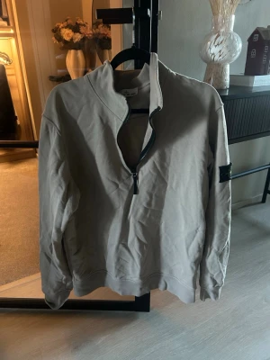 Beige half zip tröja från Stone Island - Säljer en beige långärmad tröja från Stone Island med klassisk logotyp på ärmen. Tröjan har en snygg half zip-dragkedja framtill och är gjord i mjuk bomull. Perfekt för dig som gillar stilrena och bekväma plagg med streetkänsla.
