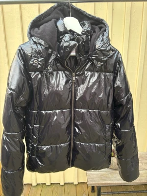 Svart pufferjacka från Nelly XS - Säljer en svart, glansig pufferjacka från Nelly i storlek XS. Jackan har huva, dragkedja framtill och är quiltad med breda paneler. Materialet är syntet och jackan har en cool, modern look som passar perfekt när det är kallt ute.