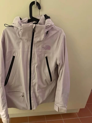 Ljuslila vindjacka från The North Face - Säljer en ljuslila vindjacka från The North Face med huva och svarta dragkedjor. Jackan har flera fickor med dragkedja och justerbara ärmslut. Perfekt för dig som gillar stilren och sportig look. Passar dig som vill ha något snyggt och funktionellt.