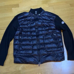 Svart pufferjacka från Moncler - Säljer en svart pufferjacka från Moncler med quiltad front i blank polyester och stickade ärmar. Jackan har dragkedja framtill, två fickor med dragkedja och en liten Moncler-logga på ärmen. Perfekt för dig som vill ha en stilren och sportig look luva finns den skickas med.