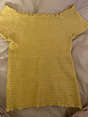 Gul smockad topp från U - Säljer en ljusgul smockad topp från U i storlek 134/140. Toppen har korta ärmar och volangkant vid hals och nederdel. Materialet är mjukt och stretchigt, perfekt för sommarens varma dagar.