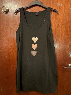 Svart linnetopp med hjärtan från SHEIN - Svart ärmlös linnetopp från SHEIN i storlek M. Framtill finns tre hjärtan i olika färgtoner: beige, ljusbrun och grå. Toppen har rund halsringning och är tillverkad i mjukt syntetmaterial. Perfekt för sommarens alla dagar.