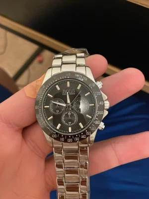 Silverfärgad Hugo Boss klocka - Stilren klocka från Hugo Boss med svart urtavla och silverfärgat armband i rostfritt stål. Klockan har tre mindre urtavlor, tydliga indexmarkeringar och en sportig bezel med siffror. Länken är robust och har Boss-loggan på spännet.