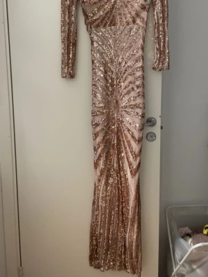 Glittrig långklänning i guld - Säljer en superglittrig långklänning i guld med coolt mönster av paljetter som strålar ut från midjan. Klänningen har långärmade och figurnära passform, perfekt för att sticka ut på festen. Materialet är syntet med massor av paljetter för extra bling.