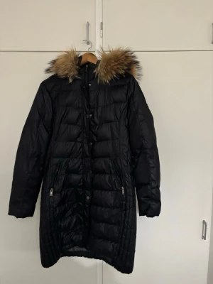 Svart lång dunjacka med päls - Säljer en svart lång dunjacka med fluffig päls runt huvan. Jackan har quiltad design, dragkedjor på fickorna och knappar framtill. Perfekt för kalla dagar och har en snygg, stilren look med mörka toner och detaljer i päls.