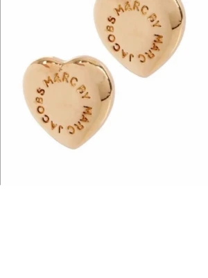 Guldiga hjärtformade örhängen Marc Jacobs - Säljer ett par snygga hjärtformade örhängen från Marc Jacobs i guldig metall. De har en stilren design med märkets logga ingraverad runt kanten. Perfekta för att ge din look lite extra glans och personlighet.