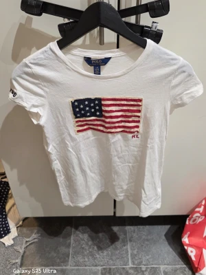 Vit t-shirt med amerikansk flagga Polo Ralph Lauren - Vit t-shirt från Polo Ralph Lauren i 100% bomull. Framsidan har en broderad amerikansk flagga med RL-detalj. Rund hals och korta ärmar, perfekt för dig som gillar klassisk och cool stil.