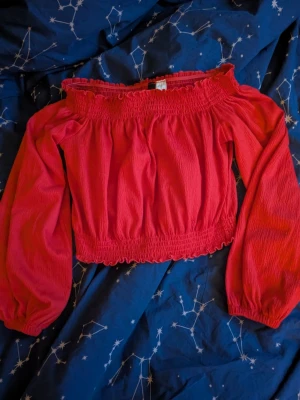 Röd offshoulder blus från H&M Divided - Säljer en röd offshoulder blus från H&M Divided i storlek S. Blusen har långa puffärmar, smock vid axlar och midja samt en croppad passform. Materialet är lätt och känns som bomull, perfekt för att ge en chill och trendig vibe.
