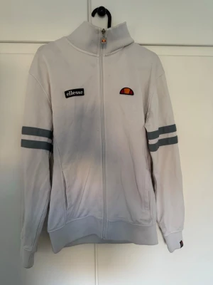 Ellesse Roma, S - Säljer en vit vindjacka från Ellesse i storlek S. Jackan har hel dragkedja, högt krage och två gråa ränder på ärmarna. Klassisk Ellesse-logga och patch på bröstet. Perfekt för dig som gillar sportig och clean stil.