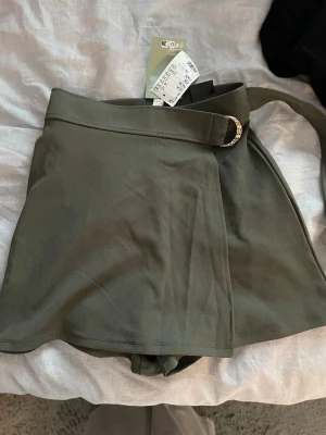 Olivgröna skorts med bälte XS - Snygga olivgröna skorts med bred midja och dekorativt bälte med metallspänne. Modellen har en kjolliknande framsida och shorts under, vilket ger en cool och bekväm look. Materialet är mjukt och lätt, perfekt för varma dagar.