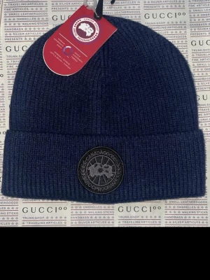 Ingen väst på bilden - Bilden visar en mörkblå ribbstickad mössa från Canada Goose med rund logotyp framtill. Ingen väst syns på bilden.